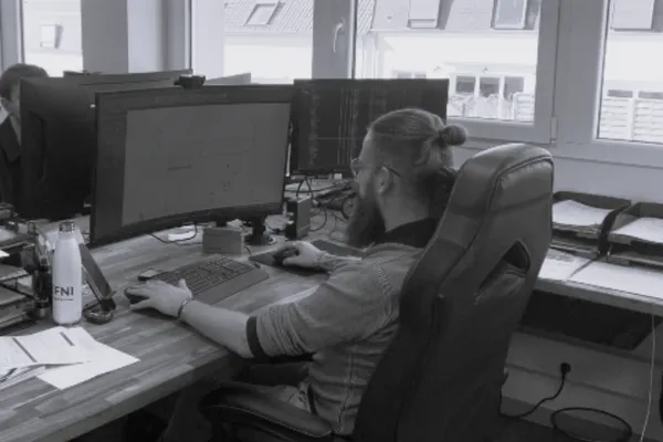 Une personne aux cheveux longs coiffés en chignon, assise à un bureau avec plusieurs écrans d'ordinateur.